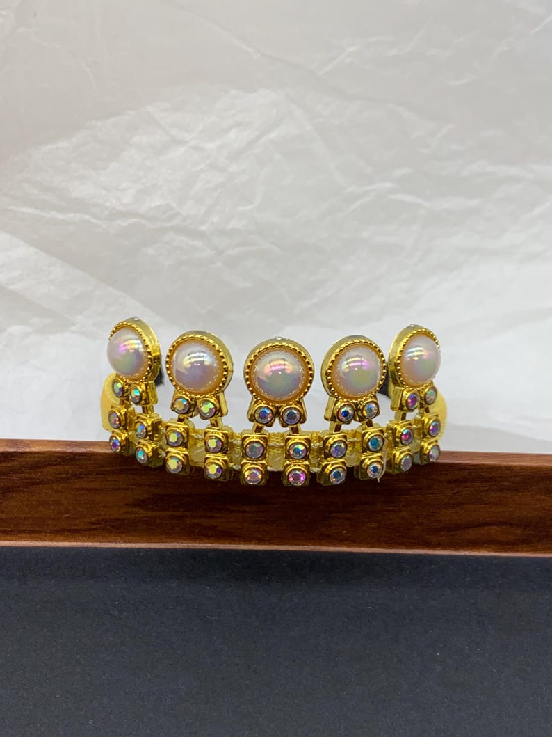 May include: A gold-toned tiara-style headband with iridescent pearl-like accents. The headband features a series of round, pearl-like stones set in gold frames, interspersed with smaller, colorful, iridescent stones. The design is symmetrical and ornate, suitable for special occasions.