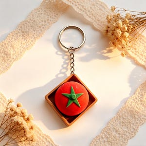 May include: A tomato-shaped keyring featuring a red tomato with a green star design, set within a brown square frame. It's attached to a silver chain and ring. The background includes lace and dried flowers.