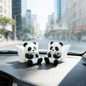 May include: Two panda bear figurines, each holding a small white device, sit on a car dashboard. The pandas are black and white, with small white pillows behind them. The background shows a city street.