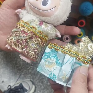 May include: A plush white bunny doll with a smiling face and two small outfits. One outfit is a red and gold embroidered skirt, and the other is a blue floral patterned skirt with gold trim. The doll is held in a person's hands.