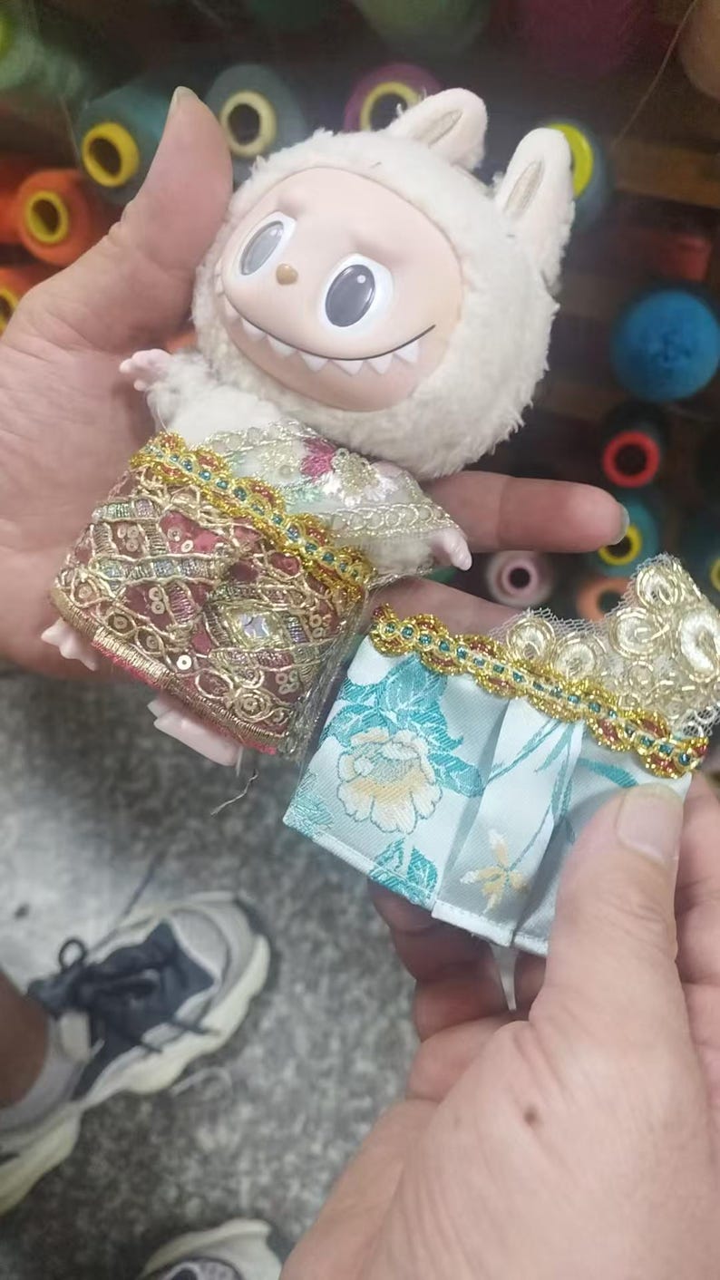 May include: A plush white bunny doll with a smiling face and two small outfits. One outfit is a red and gold embroidered skirt, and the other is a blue floral patterned skirt with gold trim. The doll is held in a person's hands.