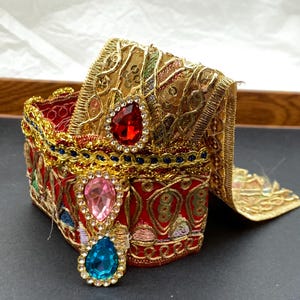 May include: A decorative red and gold box with ornate embroidery and jewel accents. The box features a red interior, gold trim, and three teardrop-shaped jewels in red, pink, and blue. The box is embellished with gold filigree and colorful embroidery.