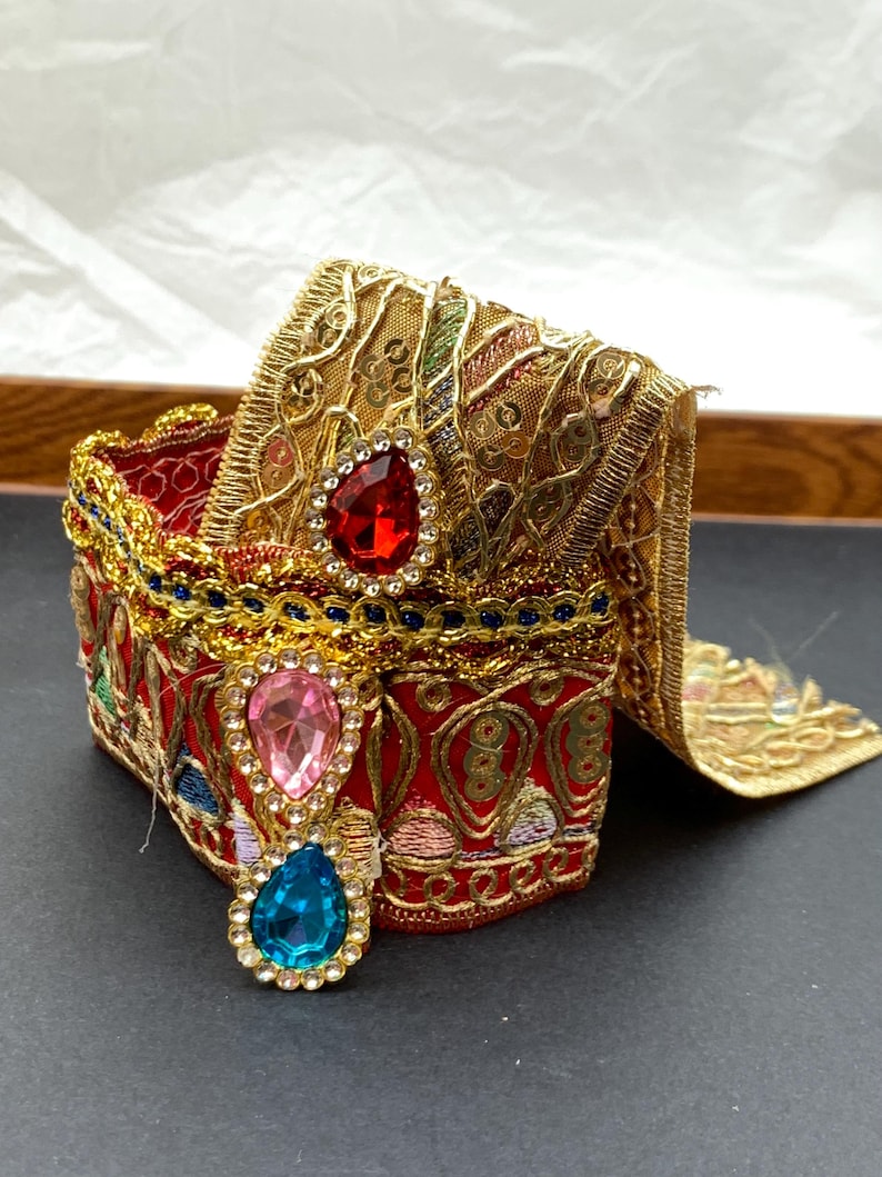 May include: A decorative red and gold box with ornate embroidery and jewel accents. The box features a red interior, gold trim, and three teardrop-shaped jewels in red, pink, and blue. The box is embellished with gold filigree and colorful embroidery.