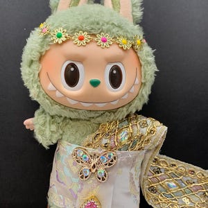 May include: A plush toy with a green furry hood and bunny ears, wearing a floral headband. The toy has a smiling face and is dressed in a white and gold outfit with a butterfly brooch and a pink jewel.
