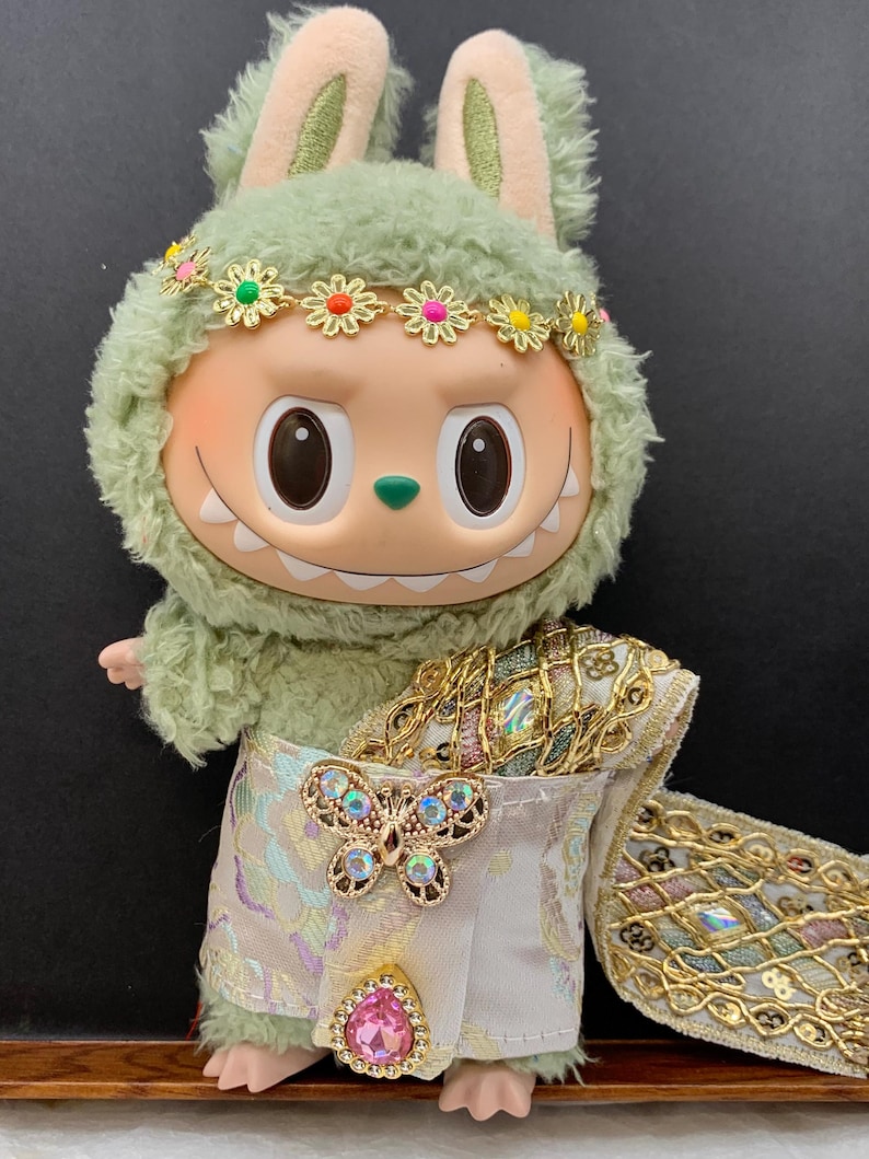 May include: A plush toy with a green furry hood and bunny ears, wearing a floral headband. The toy has a smiling face and is dressed in a white and gold outfit with a butterfly brooch and a pink jewel.