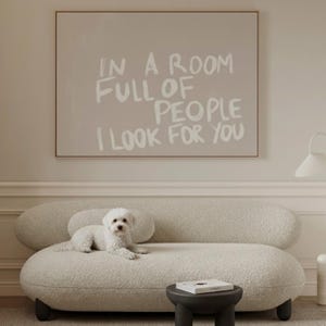 May include: A cream-coloured, curved sofa with a small, round, black side table and a white dog. A framed artwork with the text "IN A ROOM FULL OF PEOPLE I LOOK FOR YOU" hangs above the sofa.