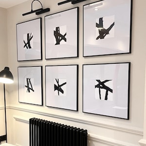 May include: Six framed abstract art prints with black frames and white matting. Each print features a unique black brushstroke design on a white background. Two black wall-mounted picture lights are above the top row of prints.
