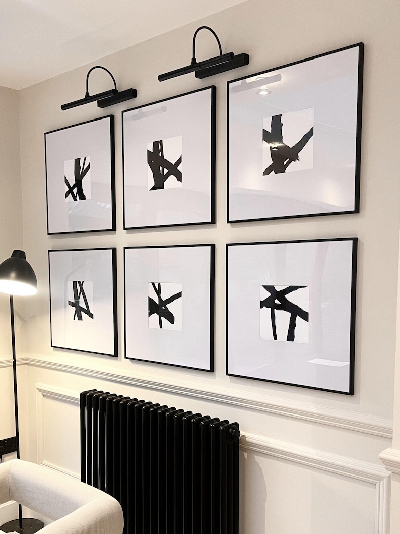 May include: Six framed abstract art prints with black frames and white matting. Each print features a unique black brushstroke design on a white background. Two black wall-mounted picture lights are above the top row of prints.