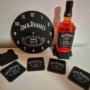 Può includere: Un orologio nero Jack Daniel's, una bottiglia di whisky con una luce rossa e sottobicchieri abbinati. L'orologio e i sottobicchieri presentano il logo Jack Daniel's in bianco. La bottiglia è esposta su un supporto nero.