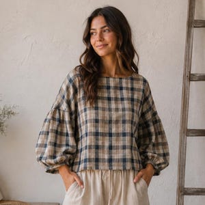 May include: A long-sleeved blouse featuring a tan and navy blue plaid pattern with voluminous sleeves and a round neckline, paired with high-waisted, wide-leg trousers in a solid light beige colour with a drawstring waist and side pockets.