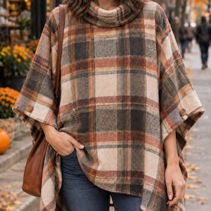 May include: A plaid poncho featuring a cowl neckline and button details on the sides. The garment displays a pattern of intersecting lines in shades of cream, brown, and burnt orange. The fabric has a textured appearance, and the design includes an asymmetrical hemline that drapes over the shoulders and torso, providing a loose fit over dark blue denim jeans.