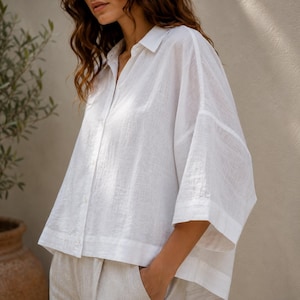 May include: A crisp white button-down shirt with a relaxed, oversized fit, featuring a classic collar, wide three-quarter length sleeves, and a straight hemline. The garment is crafted from a lightweight, semi-sheer fabric with a visible woven texture.