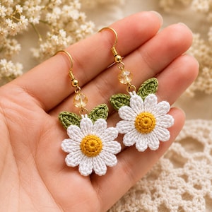 Può includere: Un paio di orecchini pendenti a forma di margherita realizzati all'uncinetto con petali bianchi e centro giallo, arricchiti da due piccole foglie verdi all'uncinetto. Ogni orecchino pende da un gancio in metallo dorato con una perlina sfaccettata.