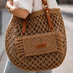 May include: A tan crochet tote bag featuring an open diamond-patterned weave, brown leather straps with gold-tone metal rings, and a rectangular front pocket with fringe detailing and a small brown leather brand label centered on the front.