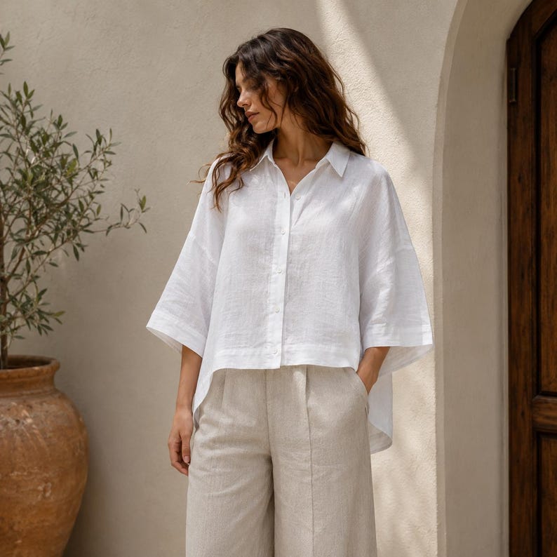 May include: A crisp white button-down shirt with a relaxed fit, featuring a classic collar, wide three-quarter length sleeves, and a high-low hemline, paired with light beige wide-leg trousers that have a visible center seam detail.