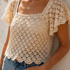 May include: A cream-colored crochet top featuring a square neckline and short, flared sleeves with a delicate shell stitch pattern. The garment has a scalloped hemline and is paired with blue denim jeans, creating a textured, open-knit appearance.