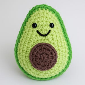 以下が含まれることがあります： 手作りのかぎ針編みのアボカドのおもちゃ。アボカドは、濃い緑色の縁と茶色の種が付いた明るい緑色です。黒い目と湾曲した黒い口で笑顔の顔をしています。おもちゃは幸せなアボカドのように見えるように設計されています。