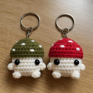 Puede incluir: Dos llaveros de setas de ganchillo hechos a mano. Uno tiene una gorra verde con lunares blancos, el otro una gorra roja con lunares blancos. Ambos tienen cuerpos blancos, ojos negros y llaveros plateados. Los llaveros son pequeños y caprichosos.