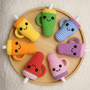 Könnte beinhalten: Eine Sammlung von sieben bunten, gehäkelten Spielzeugen in Getränkeform mit lächelnden Gesichtern. Die Spielzeuge sind auf einem runden Holztablett angeordnet. Jedes Spielzeug hat eine andere Farbe, darunter Gelb, Orange, Rot, Rosa, Lila, Blau und Grün.