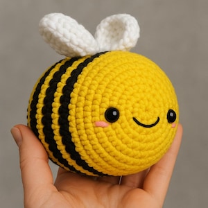 Può includere: Un'ape all'uncinetto fatta a mano con un viso sorridente, occhi neri e guance rosa. L'ape ha un corpo giallo con strisce nere e ali bianche. Questo peluche è un oggetto carino e coccoloso.