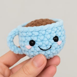 Peut inclure: Une tasse à café crochetée bleu clair avec un visage souriant, des yeux noirs et des joues roses. La tasse est remplie de fil marron, ressemblant à du café. La tasse a une anse et est faite à la main.