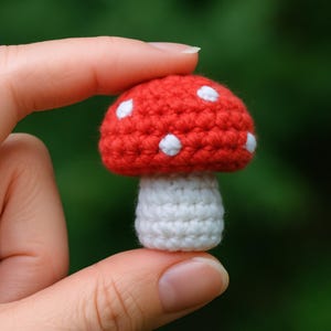 May include: A small, crocheted mushroom with a red cap and white spots. The stem is white. The mushroom is held between two fingers against a blurred green background. The handmade item is a whimsical decorative piece.