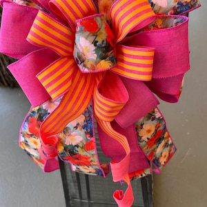 Könnte beinhalten: Eine dekorative Schleife aus einer Mischung aus einfarbigem, leuchtend pinkem Band, orange-pink gestreiftem Band und einem Blumenmusterband mit roten, orangen und weißen Blüten auf hellem Grund. Die Schleife hat mehrere Schlaufen und lange, gedrehte Enden.