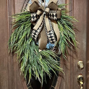 May include: A teardrop-shaped wreath featuring lush green foliage and a large decorative bow. The bow is crafted from tan hessian ribbon and a black and white ribbon with a diamond pattern. The wreath is suspended by a metal hook.