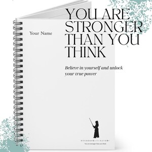 以下が含まれることがあります： 「YOU ARE STRONGER THAN YOU THINK」と「Believe in yourself and unlock your true power」の文字が書かれた白いリングノート。表紙には「Your Name」の文字と人物のシルエットも描かれています。Spirit and Strength Collectionの一部です。