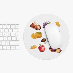 May include: White circular mouse mat featuring a watercolour illustration of autumnal produce. The design includes a red apple, persimmons, grapes, chestnuts, mushrooms, and sweet potatoes. A white computer mouse rests on the mat.