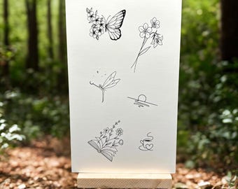 May include: A white card on a small wooden easel displays delicate black line art. The illustrations include a butterfly with flowers, floral sprigs, a fairy, a sunset, an open book with flowers, and a cup of coffee. The background is a blurred forest.