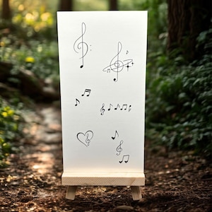 Op de afbeelding: Een wit canvas op een kleine houten ezel toont verschillende zwarte muzieknoten en symbolen, waaronder G-sleutels, achtste noten en een hartvormige sleutel. De achtergrond is een vage buitenscène met groen gebladerte.