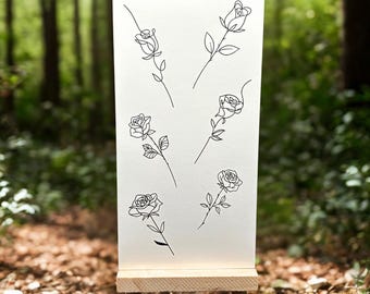 Könnte beinhalten: Eine weiße Karte auf einer kleinen Holzstaffelei zeigt sechs schwarze Strichzeichnungen von Rosen und Stielen. Das Kunstwerk befindet sich vor einem verschwommenen Hintergrund eines Waldes mit braunen Blättern und grünen Bäumen.