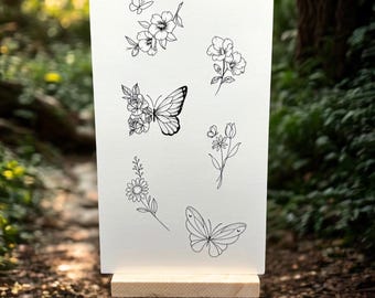 May include: A white card on a small wooden easel displays delicate black line drawings of butterflies and flowers. The designs include a butterfly with floral wings, individual flowers, and other butterfly illustrations. The background is a blurred forest scene.