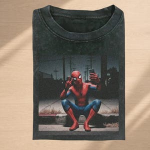 May include: A black acid-wash t-shirt featuring a graphic print of Spider-Man in a red and blue suit, crouching on a curb at night while taking a selfie with a smartphone. The character is making a peace sign with one hand and holding the phone.