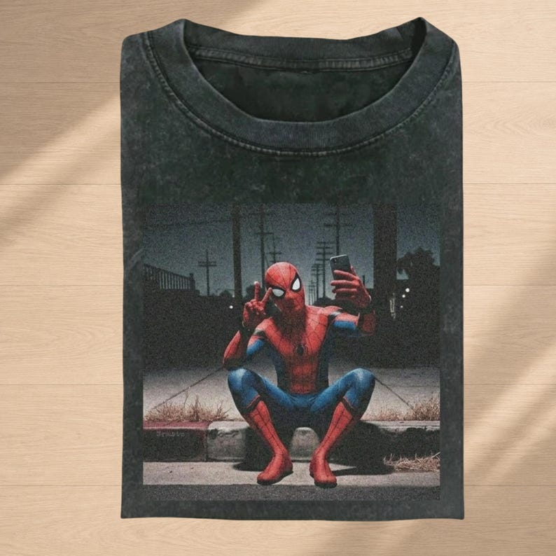 May include: A black acid-wash t-shirt featuring a graphic print of Spider-Man in a red and blue suit, crouching on a curb at night while taking a selfie with a smartphone. The character is making a peace sign with one hand and holding the phone.