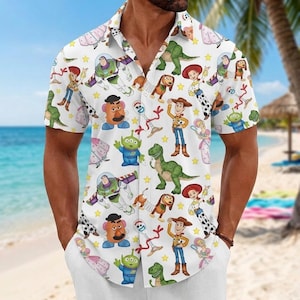 Op de afbeelding: Een wit overhemd met korte mouwen en knoopsluiting, voorzien van een all-over print van diverse stripfiguren, waaronder een space ranger, een cowboy, een cowgirl, een groene dinosaurus, een aardappelvormig figuur en gele sterren.
