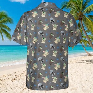 May include: Gray short-sleeved button-down shirt with a repeating pattern of a green alien character, a helmet, and a dark brown bird-like creature. The shirt is on a sandy beach with palm trees and a blue ocean in the background.
