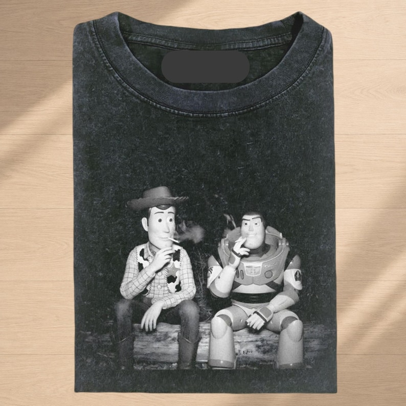 Puede incluir: Una camiseta negra con efecto lavado al &aacute;cido que presenta un estampado gr&aacute;fico en blanco y negro de los personajes Woody y Buzz Lightyear sentados juntos en un tronco, cada uno sosteniendo un cigarrillo, con un fondo de bosque oscuro.