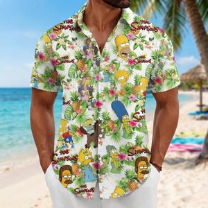 Könnte beinhalten: Ein kurzärmeliges Hemd mit Knopfleiste und einem Allover-Druck der Simpsons-Figuren, darunter Homer, Marge, Bart, Lisa und Maggie, durchsetzt mit tropischen Ananas, grünen Palmblättern und rosa Hibiskusblüten auf einem weißen Hintergrund.