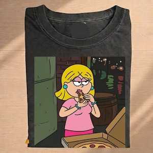May include: A charcoal gray t-shirt featuring a graphic illustration of a blonde cartoon character wearing a pink shirt and blue earrings, eating a slice of pizza. The background shows a brick wall, a wooden barrel, and an open pizza box.