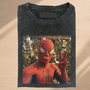 Puede incluir: Una camiseta gris carb&oacute;n de manga corta con cuello redondo y acabado lavado vintage. El centro presenta un estampado gr&aacute;fico de un superh&eacute;roe enmascarado rojo y azul haciendo el signo de la paz frente a un paisaje urbano nocturno.