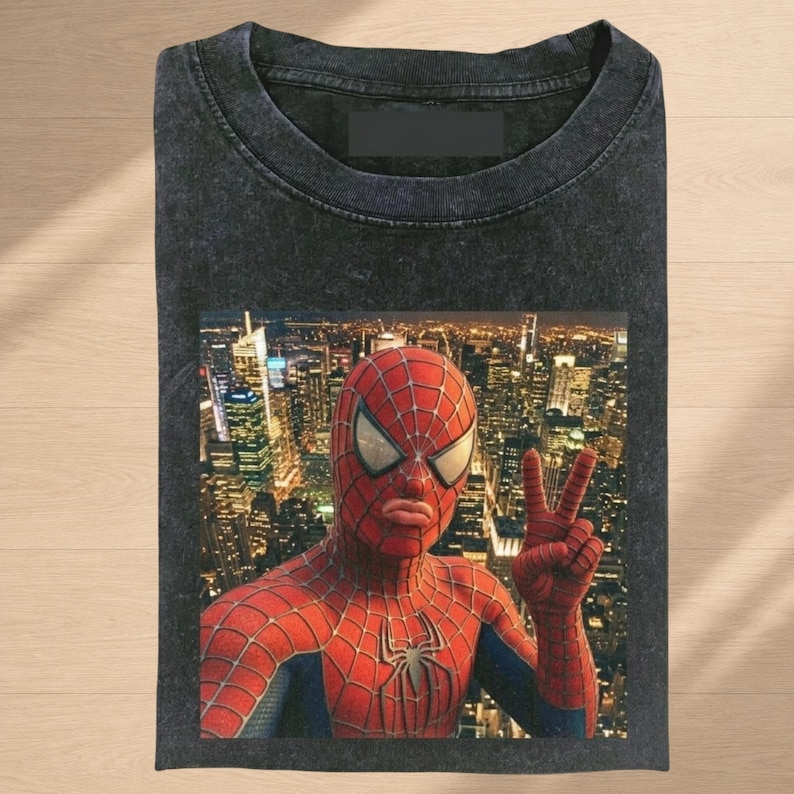 Puede incluir: Una camiseta gris carb&oacute;n de manga corta con cuello redondo y acabado lavado vintage. El centro presenta un estampado gr&aacute;fico de un superh&eacute;roe enmascarado rojo y azul haciendo el signo de la paz frente a un paisaje urbano nocturno.