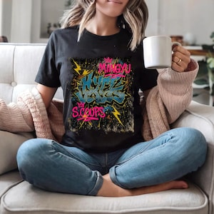 May include: Black t-shirt with a graffiti-style graphic. The design features the words "Hype Voices" in blue and "Mingyu" and "S.Coups" in pink, with yellow lightning bolts and a crown. The shirt is short-sleeved.