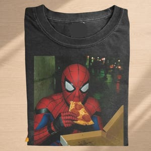 May include: A black crew neck t-shirt featuring a graphic print of a red and blue costumed character wearing a mask and eating a slice of pepperoni pizza from an open cardboard box. The shirt has a dark, washed-out fabric texture and a round neckline.