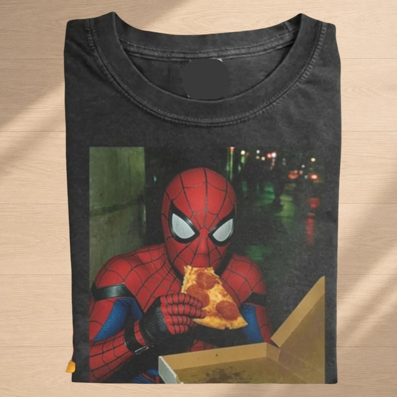 May include: A black crew neck t-shirt featuring a graphic print of a red and blue costumed character wearing a mask and eating a slice of pepperoni pizza from an open cardboard box. The shirt has a dark, washed-out fabric texture and a round neckline.