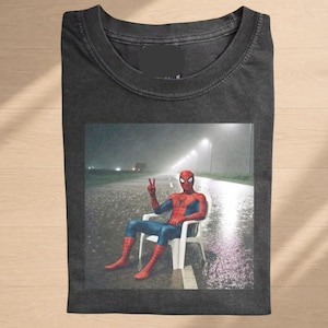 May include: A charcoal gray crew neck t-shirt featuring a graphic print of the character Spider-Man sitting on a white plastic chair in the middle of a rainy, dark road at night, making a peace sign with one hand while streetlights glow in the distance.
