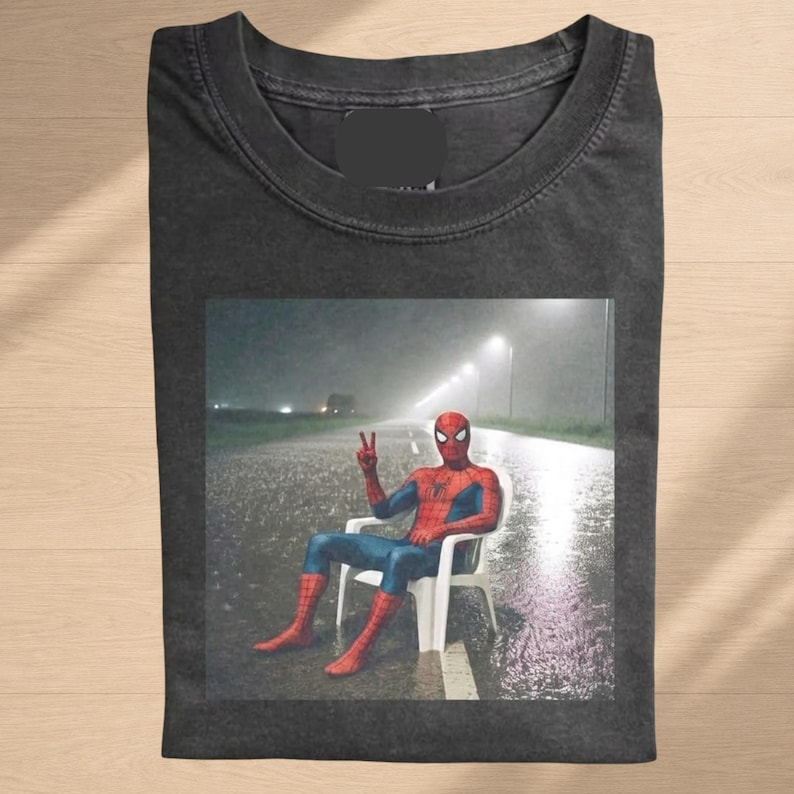 May include: A charcoal gray crew neck t-shirt featuring a graphic print of the character Spider-Man sitting on a white plastic chair in the middle of a rainy, dark road at night, making a peace sign with one hand while streetlights glow in the distance.