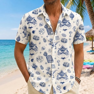 May include: Short-sleeved button-down shirt featuring a repeating pattern of a cat wearing a crown, dinosaurs, treasure chests, and other whimsical illustrations in blue on a cream background. The shirt is worn by a person on a beach.