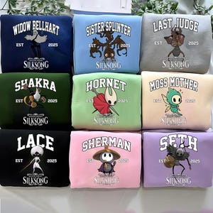May include: Nine colorful sweatshirts with various character designs and text from the video game Hollow Knight: Silksong. Each sweatshirt features a different character name, the year 2025, and the game's title. The sweatshirts are in various colors.